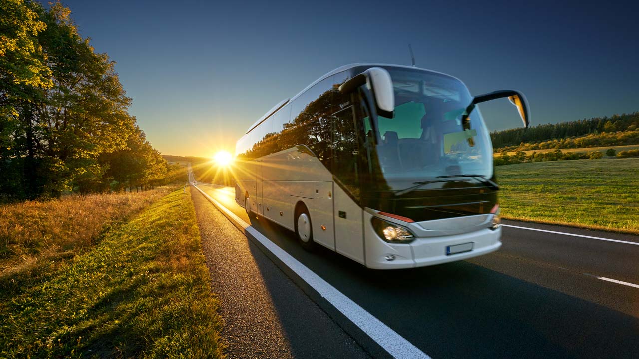 Illustrasjon av buss som kjører langs en veg, med lav sol i bakgrunnen. Høstlandskap.